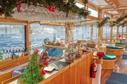 The image shows a festively decorated bar onboard a boat, adorned with Christmas decorations and a view of the water outside.