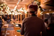 A person wearing a Santa hat is facing away from the camera, overlooking a festive and warmly-lit interior adorned with holiday decorations.