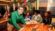 A waiter is presenting a tray of food to a group of smiling guests at a festively decorated venue.