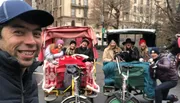 A joyful group of people are taking pedicab rides in what appears to be a city park setting, smiling and posing for a group selfie.