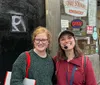 Two people are smiling in front of Yonah Schimmels Knishery which has various signs indicating social distancing rules and business hours