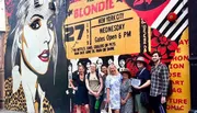 A group of people are smiling for a photo in front of a colorful wall mural featuring an advertisement for a Blondie concert in New York City.
