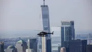 A helicopter is flying in the foreground with a backdrop of skyscrapers that includes a very tall building with a flat top, possibly in a large city's downtown area.