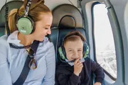 A woman and a young child are smiling and wearing headphones while sitting inside a helicopter, with a cityscape visible through the window.