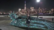 A person is posing next to a sculpture of a giant head resembling the Statue of Liberty with the illuminated New York City skyline in the background at night.