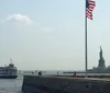 An American flag flies in the foreground with the Statue of Liberty in the distance while a ferry carries passengers nearby