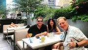 Three people are smiling for the camera while seated at an outdoor table with drinks, in a setting that looks like a rooftop or patio bar with lush greenery and city buildings in the background.