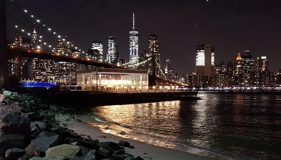 The image captures a nighttime view of the illuminated Brooklyn Bridge and the New York City skyline reflected in the East River.