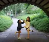 Two people are sharing a playful moment under a brick archway in a lush green park