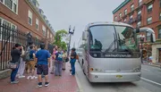 A group of tourists is gathered on a sidewalk about to board or disembarking from a tour bus on a city street.