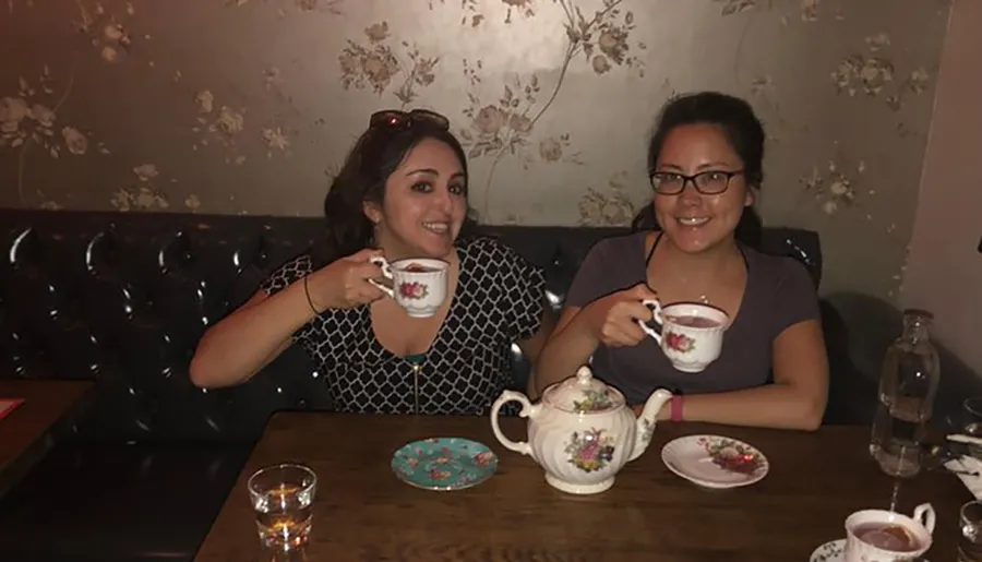 Two women are smiling and holding teacups, sitting at a table with a teapot and a glass of water.