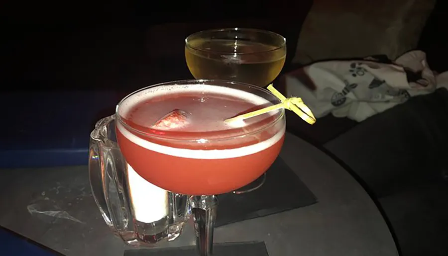 The image shows two cocktails on a table in a dimly lit setting, with a frothy reddish drink in the foreground garnished with a wedge on a cocktail stick and a clear yellowish drink in the background, alongside a person whose lap is partially visible.