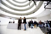 The image shows the interior of a museum with a spiral walkway, where numerous visitors are either walking around or engaged in conversation.