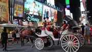 A horse-drawn carriage is navigating through a bustling Times Square lit by colorful billboards and signs at dusk.