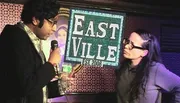 A man holding a microphone speaks to a woman on a stage with a sign that reads 