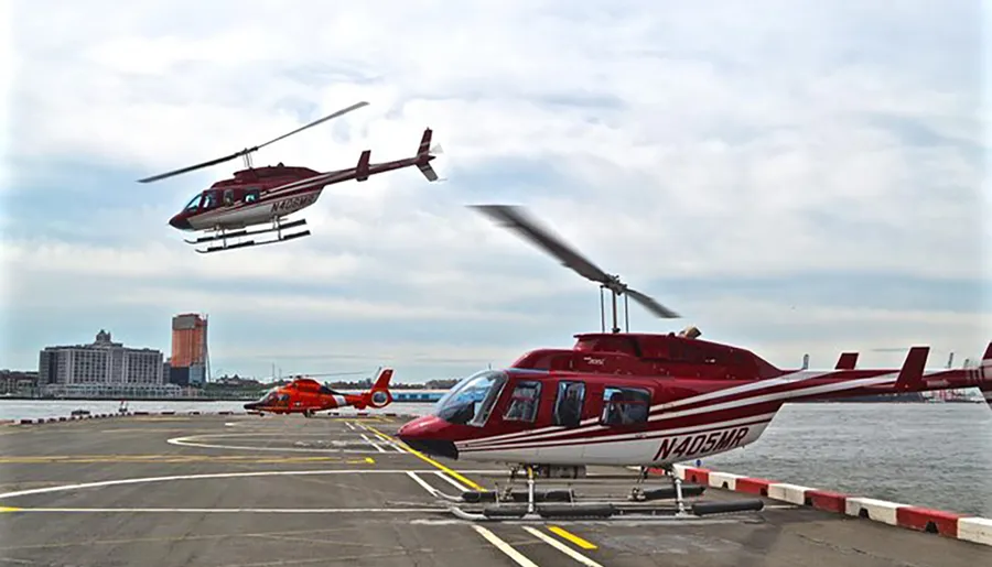 Three helicopters, one in air and two parked, are at a helipad by the water with city buildings in the background.