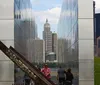 The image shows visitors at a reflective memorial with a weathered metal beam in the foreground and a cityscape in the background