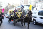 A horse-drawn carriage adorned with yellow and pink plumes and decorations is trotting through an urban street with passengers and a driver onboard, amidst modern vehicular traffic.