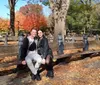 Two people are smiling for a photo on a sunny day sitting on a park bench amidst fallen autumn leaves with colorful trees in the background