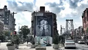 This image features a large building mural of a child's face with an urban street scene and the Williamsburg Bridge in the background.