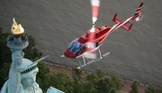 A red helicopter is flying near the torch of the Statue of Liberty against a background of water.