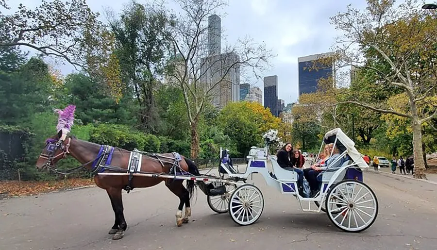 A horse-drawn carriage with passengers is traversing a path in a park with a city skyline in the background.