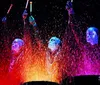 Three performers with blue-painted faces and speckled black shirts appear illuminated under a dark background