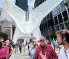 This image captures a bustling street scene in front of the distinctive wing-like structure of the Oculus in New York City with pedestrians engaging in various activities under a blue sky peppered with clouds