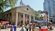 People are gathered around Quincy Market on a sunny day, with street vendors and an American flag adorning the facade.