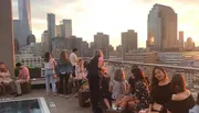 A group of people are enjoying a rooftop gathering during sunset with a view of a city skyline.