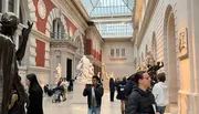 Visitors explore an art gallery featuring classical sculptures and architectural designs, with natural light coming in from a glass ceiling.
