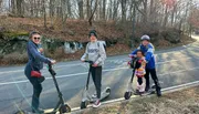 Four individuals are smiling and posing with electric scooters on a roadside, with bare trees and a clear sky in the background.