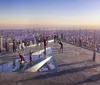 This image shows people enjoying a panoramic view of a city skyline from a high observation deck with glass floor panels