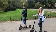 Two people are riding electric scooters along a path with greenery in the background.