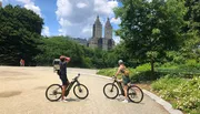 Two cyclists take a break in a park with lush greenery and a backdrop of tall buildings under a clear sky.