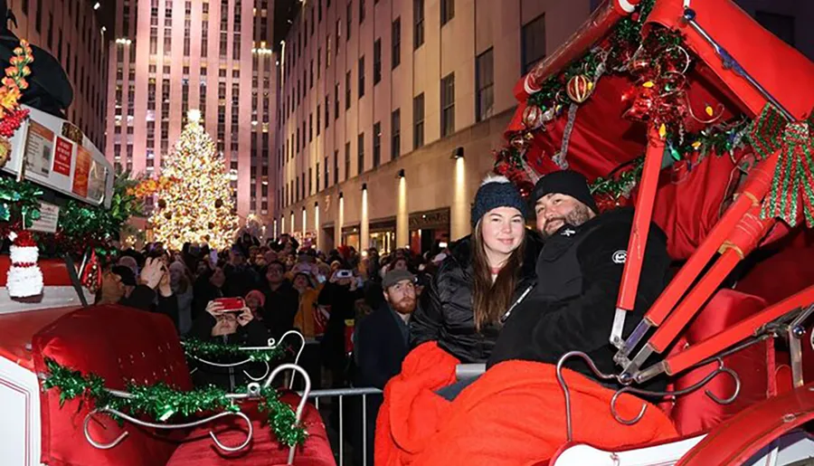 A couple poses for a photo in a decorated horse-drawn carriage with a festive crowd and a large Christmas tree in the background.