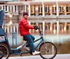 A person in a red jacket and purple hat is riding a pedicab by a reflective body of water with a building illuminated by warm lights in the background