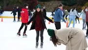 A group of people is enjoying their time on an ice skating rink, with a smiling woman in the foreground showing off her balance while another person appears to be bending down or falling in the foreground.