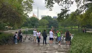 Visitors are enjoying a cloudy day by a pond in a lush park with the city skyline in the background.