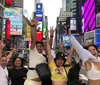 A group of joyful people are posing with raised arms in a bustling Times Square surrounded by bright advertisements and city life