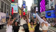 A group of joyful people are posing with raised arms in a bustling Times Square, surrounded by bright advertisements and city life.