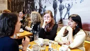 A group of people appears engaged in conversation at a table in a café or restaurant with a large historical mural on the wall behind them.
