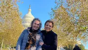 Two people are smiling for a photo in front of the United States Capitol building with autumn foliage in the background.