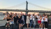 A joyful group of people poses for a photo in front of a bridge at dusk, with several individuals making peace signs or embracing each other.