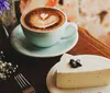 A cup of latte with heart-shaped latte art is served beside a slice of cheesecake on a wooden table accompanied by a small bouquet of flowers and a fork