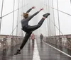 A person performs a high kick on a foggy bridge while pedestrians walk in the background