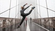 A person performs a high kick on a foggy bridge while pedestrians walk in the background.
