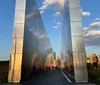 The image shows a modern reflective monument pathway with people walking through it overlooking a city skyline in the background during sunset