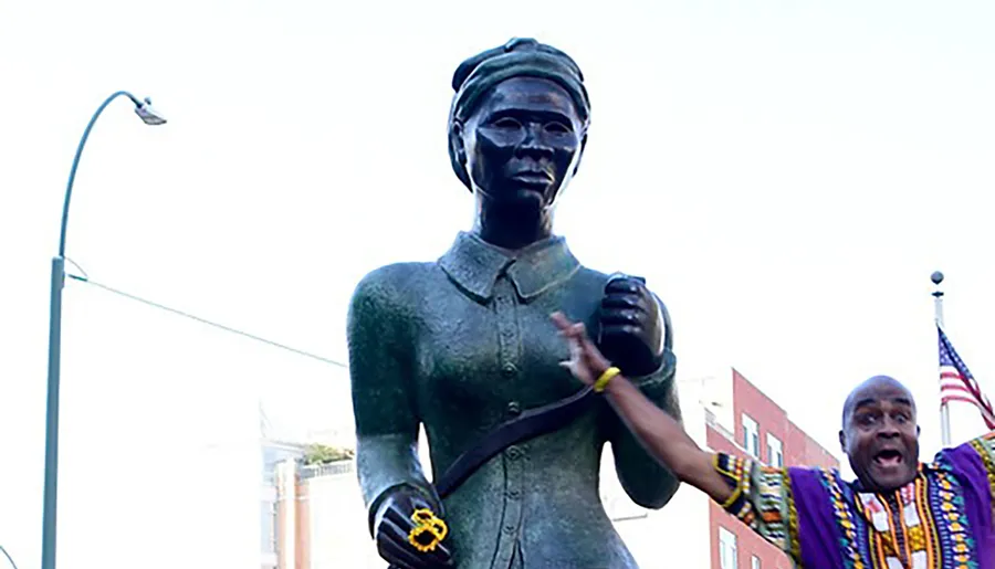 A person in colorful attire is gesturing towards a bronze statue of a standing woman.