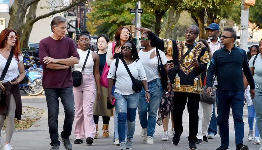 A diverse group of people appears to be walking together on a city street, possibly on a guided tour as one individual gestures toward something of interest.